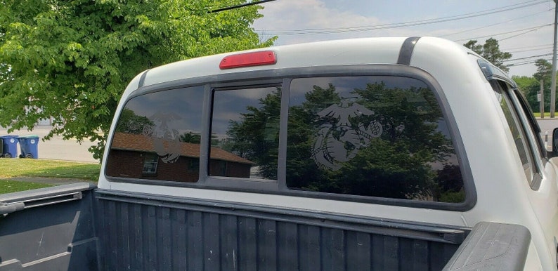 May include: A white pickup truck with a tinted rear window. The window has a black and white decal of the United States Marine Corps emblem.