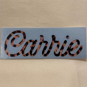 May include: A light blue background with the name "Carrie" written in a cursive font. The letters are filled with a leopard print pattern.