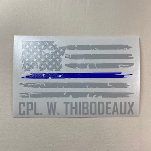 May include: A distressed American flag with a thin blue line running horizontally across the middle. The flag is printed on a white background with the text "CPL. W. THIBODEAUX" printed below the flag.