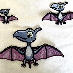 Op de afbeelding: Drie geborduurde patches met een cartoon pterodactyl ontwerp in verschillende maten. Elke dinosaurus heeft een grijze kop en lichaam, een witte buik en paarse vleugels met zwarte omlijningen. De wezens hebben grote groene ogen en vliegen.