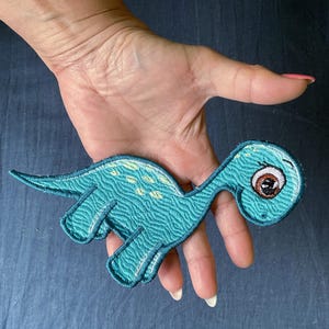 Op de afbeelding: Een turquoise geborduurde dinosaurus-patch met een lange nek, een ronde kop met een groot bruin oog en lichtgele vlekken op de rug. De patch heeft een textuurrijk oppervlak en een donker turquoise gestikte rand langs de hele vorm.