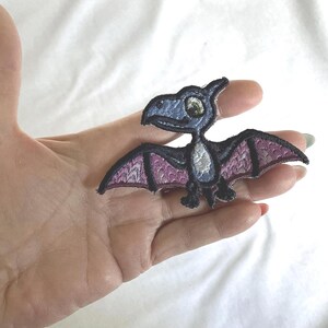 Op de afbeelding: Een geborduurde patch met een gestileerde pterodactyl met een donkerblauwe kop en lichaam, een lichtgrijze borst en vleugels met een textuur van paarse en zwarte patronen. De dinosaurusfiguur heeft een groot oog en een puntige snavel.