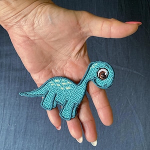 Op de afbeelding: Een geborduurde patch in de vorm van een dinosaurus met een lange nek in tinten blauw en groenblauw. De dinosaurus heeft een groot bruin oog, een glimlachende mond en lichtgele vlekken op de rug. Het borduurwerk heeft een textuur.