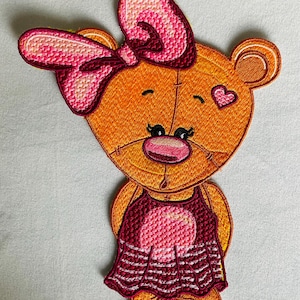 Op de afbeelding: Een geborduurde patch met een oranje beerfiguur met een roze strik op het hoofd, een klein roze hartje op de wang en een donkerrode jurk met een roze cirkelvormig patroon op de borst en witte horizontale strepen op de zoom van de rok.
