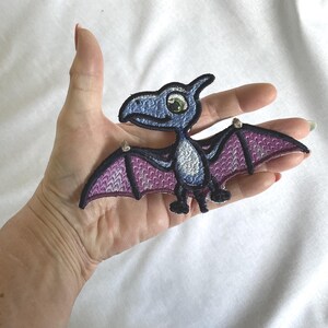 Op de afbeelding: Een geborduurde patch met een cartoon-pterodactyl met een blauwe kop en lichaam, een witte borst en paarse vleugels met zwarte omlijningen. Het wezen heeft een groot groen oog en een kleine snavel, uitgevoerd in een platte borduurstijl.
