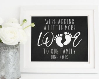 May include: A white framed chalkboard sign with black text that reads "We're adding a little more love to our family June 2019". The sign has a pair of white baby footprints in the middle of the word "love".