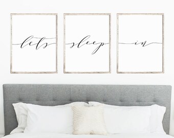 May include: Three framed wall art pieces with the words "lets", "sleep", and "in" in elegant cursive script. The frames have a light wood finish. The art hangs above a bed with a gray headboard and white bedding.