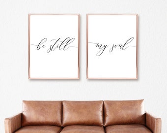 May include: Two framed prints with the text "be still" and "my soul" in a cursive font. The prints are hung on a white wall above a brown leather couch.