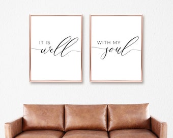 May include: Two framed wall art prints with the words "IT IS well" and "WITH MY soul" in elegant black script. The frames have a rose gold finish, and the prints are displayed above a brown leather sofa.