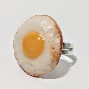 May include: A silver ring featuring a fried egg design. The egg is white with a yellow yolk and a brown, slightly crispy edge.
