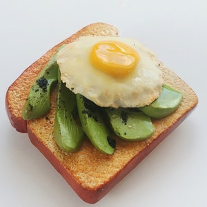 Puede incluir: Una rebanada de pan tostado cubierta con rodajas de aguacate y un huevo frito. El aguacate est&aacute; dispuesto en un patr&oacute;n circular alrededor del huevo, que tiene una yema de huevo amarilla brillante.