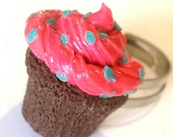 May include: A pink cupcake with blue sprinkles on top, made of polymer clay, is attached to a silver ring. The cupcake is a miniature replica of a real cupcake.