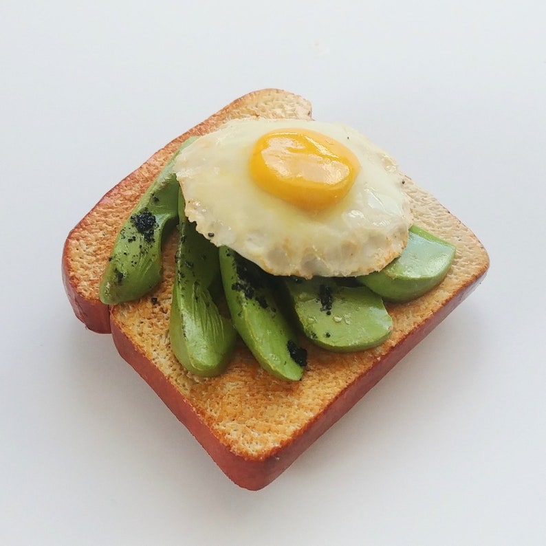 Puede incluir: Una rebanada de pan tostado cubierta con rodajas de aguacate y un huevo frito. El aguacate est&aacute; dispuesto en un patr&oacute;n circular alrededor del huevo, que tiene una yema de huevo amarilla brillante.