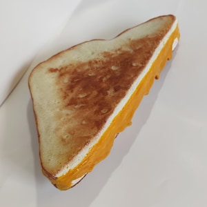 May include: A single slice of a toasted cheese sandwich. The bread is a golden brown colour, with melted orange cheese spilling out. The sandwich is on a white surface.