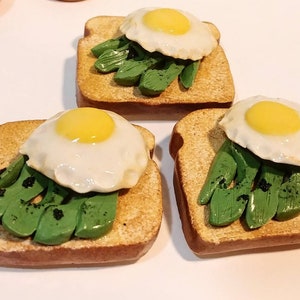 Puede incluir: Tres tostadas de aguacate en miniatura con huevos fritos. Cada tostada est&aacute; cubierta con rodajas de aguacate verde y un huevo frito con una yema de color amarillo brillante.