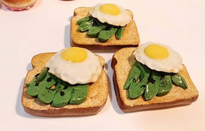 Puede incluir: Tres tostadas de aguacate en miniatura con huevos fritos. Cada tostada est&aacute; cubierta con rodajas de aguacate verde y un huevo frito con una yema de color amarillo brillante.