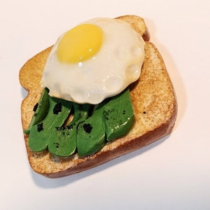 Puede incluir: Una escultura de arcilla en miniatura de una tostada cubierta con aguacate, un huevo frito y semillas de s&eacute;samo negras.
