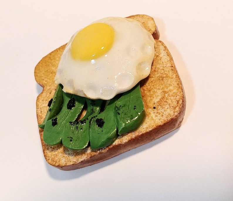 Puede incluir: Una escultura de arcilla en miniatura de una tostada cubierta con aguacate, un huevo frito y semillas de s&eacute;samo negras.
