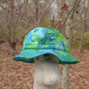 May include: A green, blue, and white tie-dye bucket hat sits on top of a gray concrete rabbit statue. The hat has a wide brim and is made of a soft, lightweight fabric.