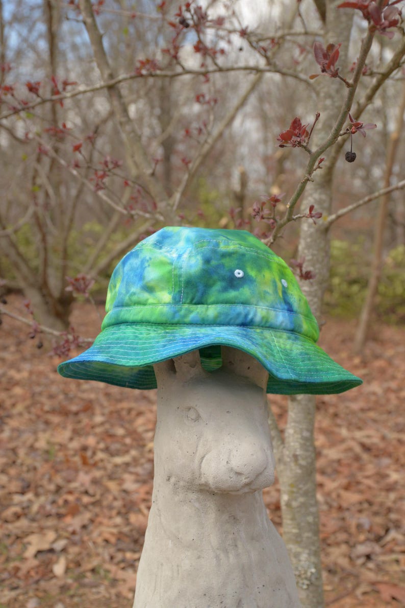 May include: A green, blue, and white tie-dye bucket hat sits on top of a gray concrete rabbit statue. The hat has a wide brim and is made of a soft, lightweight fabric.