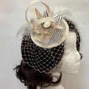Può includere: Un fascinator beige con base circolare, un nastro ad anello e piume bianche con strisce nere. Un bottone ovale bianco è posizionato al centro della composizione floreale, completato da un velo a rete bianco che copre la parte anteriore.
