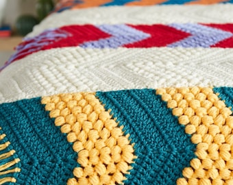 May include: A colorful crocheted throw blanket with a geometric pattern. The blanket features stripes of red, white, blue, yellow, and teal.