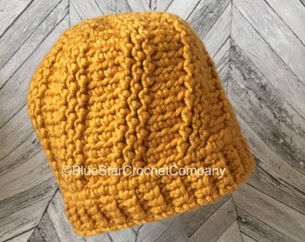 May include: A mustard yellow crocheted beanie hat. The hat is made with a textured stitch pattern. The hat is sitting on a wooden surface.