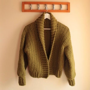 May include: A green crocheted cardigan with a shawl collar and long sleeves hanging on a wooden rack.