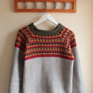 May include: A handmade crochet sweater in shades of grey, red, yellow, and olive green hangs on a white hanger. The sweater features a patterned yoke and cuffs, with a solid grey body and sleeves. A wooden coat rack is in the background.