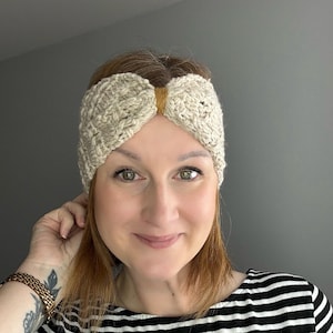 May include: A beige crocheted headband with a textured design. The headband features a brown band in the centre. The person in the image is wearing a black and white striped top.
