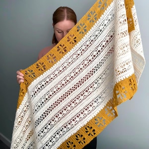 Può includere: Uno scialle all'uncinetto color crema con un bordo giallo senape con ritagli di fiori. Lo scialle ha un motivo a righe con dettagli traforati. Lo scialle fatto a mano è tenuto in mano, mostrando il suo design e la sua consistenza.