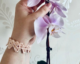 May include: A delicate pink crocheted bracelet with a floral design. The bracelet is worn on a person's wrist, and a pink orchid is held in the same hand.