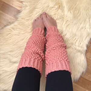Puede incluir: Un par de polainas de crochet rosa con un patrón de punto texturizado. Las polainas se llevan sobre unos leggings negros.