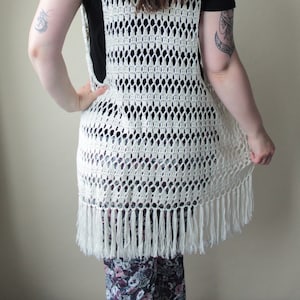 Puede incluir: Un chaleco de crochet blanco con un dobladillo de flecos. El chaleco está hecho con un delicado patrón de punto abierto.