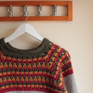 May include: A handmade crochet sweater in grey, olive green, red, and orange. The yoke features a geometric pattern in red, orange, and olive green. The sweater is hanging on a white hanger from a wooden coat rack.