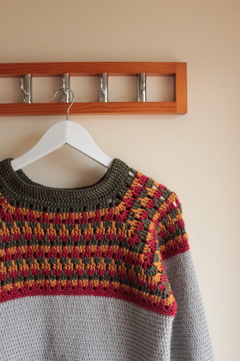 May include: A handmade crochet sweater in grey, olive green, red, and orange. The yoke features a geometric pattern in red, orange, and olive green. The sweater is hanging on a white hanger from a wooden coat rack.