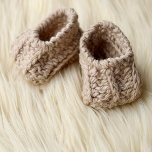 a pair of crochet baby booties in cream color on a plain white background with textured stitches made from a flat rectangle suitable for newborn baby boy or girl for baby shower gift as dyi baby shoes