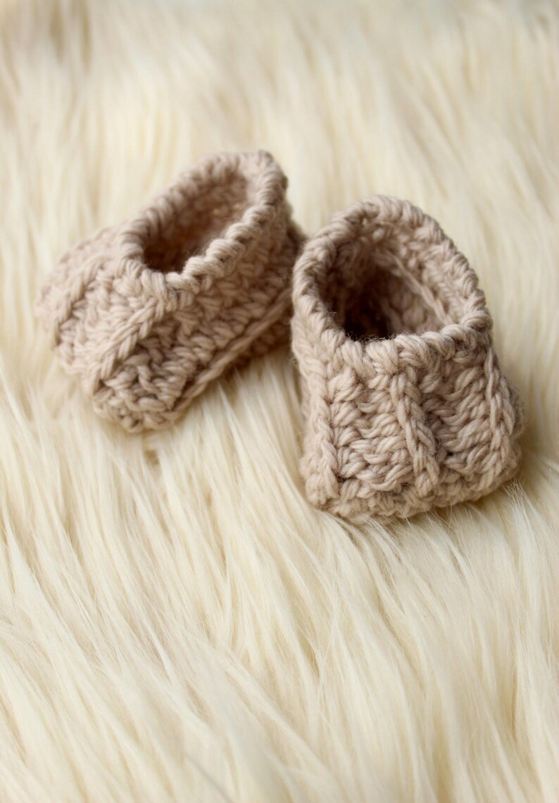 a pair of crochet baby booties in cream color on a plain white background with textured stitches made from a flat rectangle suitable for newborn baby boy or girl for baby shower gift as dyi baby shoes