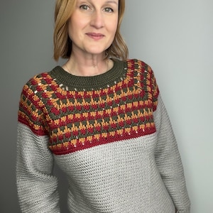 May include: A grey crochet sweater with a patterned yoke in red, orange, and green. The sweater has long sleeves and a ribbed hem. The neckline is olive green.