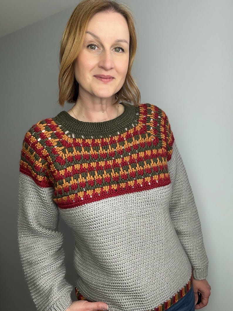May include: A grey crochet sweater with a patterned yoke in red, orange, and green. The sweater has long sleeves and a ribbed hem. The neckline is olive green.