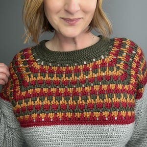May include: A handmade crochet sweater with a colour block design. The top yoke has a geometric pattern in red, orange, and green. The neckline is olive green, and the body is grey. The sweater is a cosy, handmade garment.