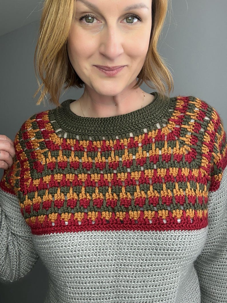 May include: A handmade crochet sweater with a colour block design. The top yoke has a geometric pattern in red, orange, and green. The neckline is olive green, and the body is grey. The sweater is a cosy, handmade garment.