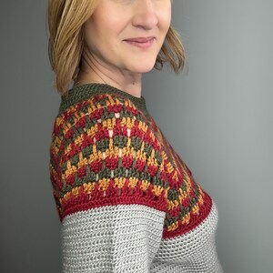 May include: A grey crochet sweater with a colourful yoke. The yoke features a geometric pattern in red, gold, and olive green. The sweater has a crew neck and is made of a soft, textured yarn.