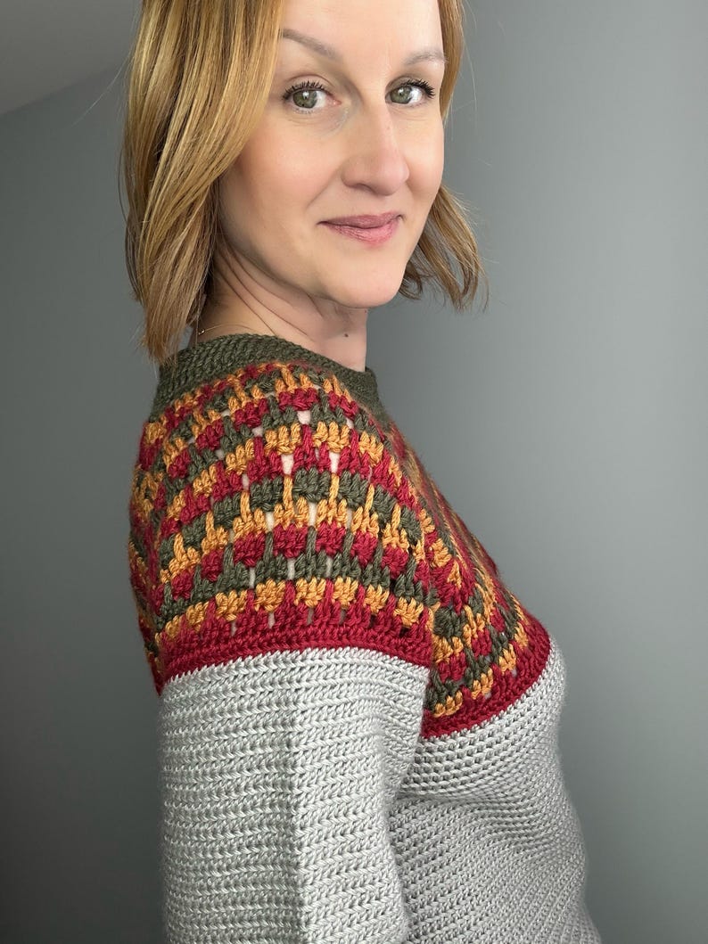 May include: A grey crochet sweater with a colourful yoke. The yoke features a geometric pattern in red, gold, and olive green. The sweater has a crew neck and is made of a soft, textured yarn.