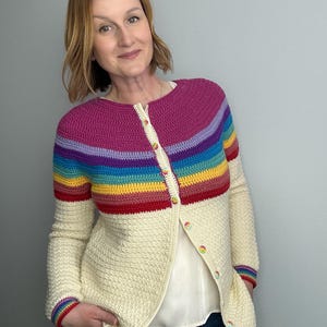 May include: A cream-colored, crocheted cardigan with a rainbow striped yoke and sleeve cuffs. The cardigan has a textured stitch pattern and is buttoned up the front with colorful buttons. The sleeves are long, and the overall style is casual.