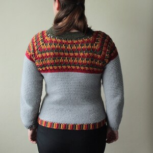 May include: A grey crochet sweater with long sleeves. The yoke features a geometric pattern in red, orange, and olive green. The bottom hem also has a striped pattern in the same colours. The sweater is worn by a person.