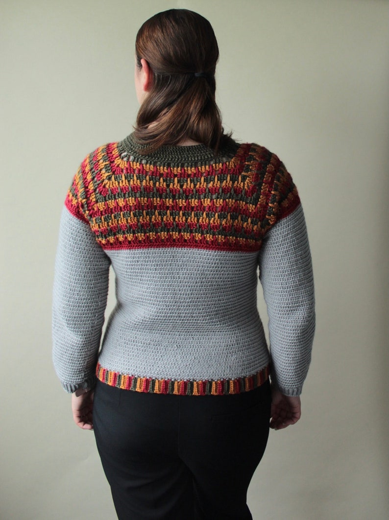 May include: A grey crochet sweater with long sleeves. The yoke features a geometric pattern in red, orange, and olive green. The bottom hem also has a striped pattern in the same colours. The sweater is worn by a person.