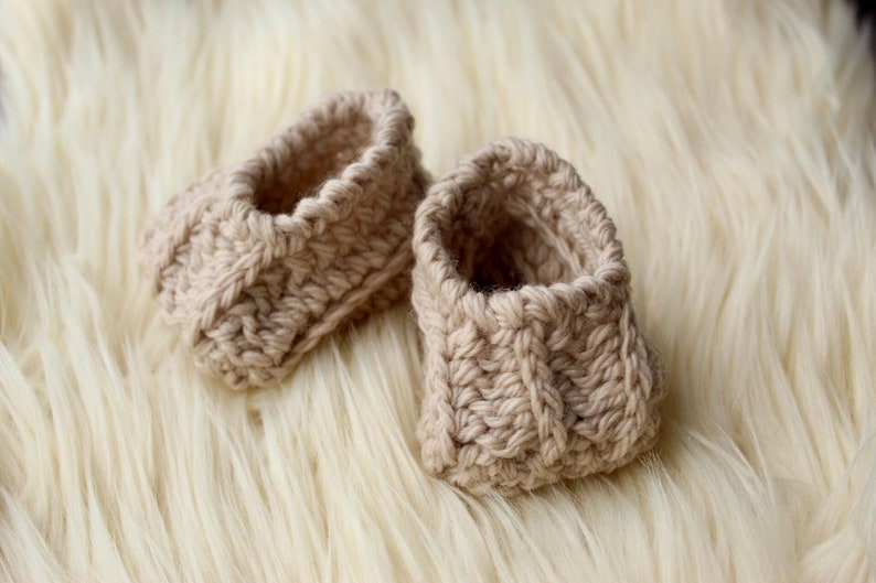 a pair of crochet baby booties in cream color on a plain white background with textured stitches made from a flat rectangle suitable for newborn baby boy or girl for baby shower gift as dyi baby shoes