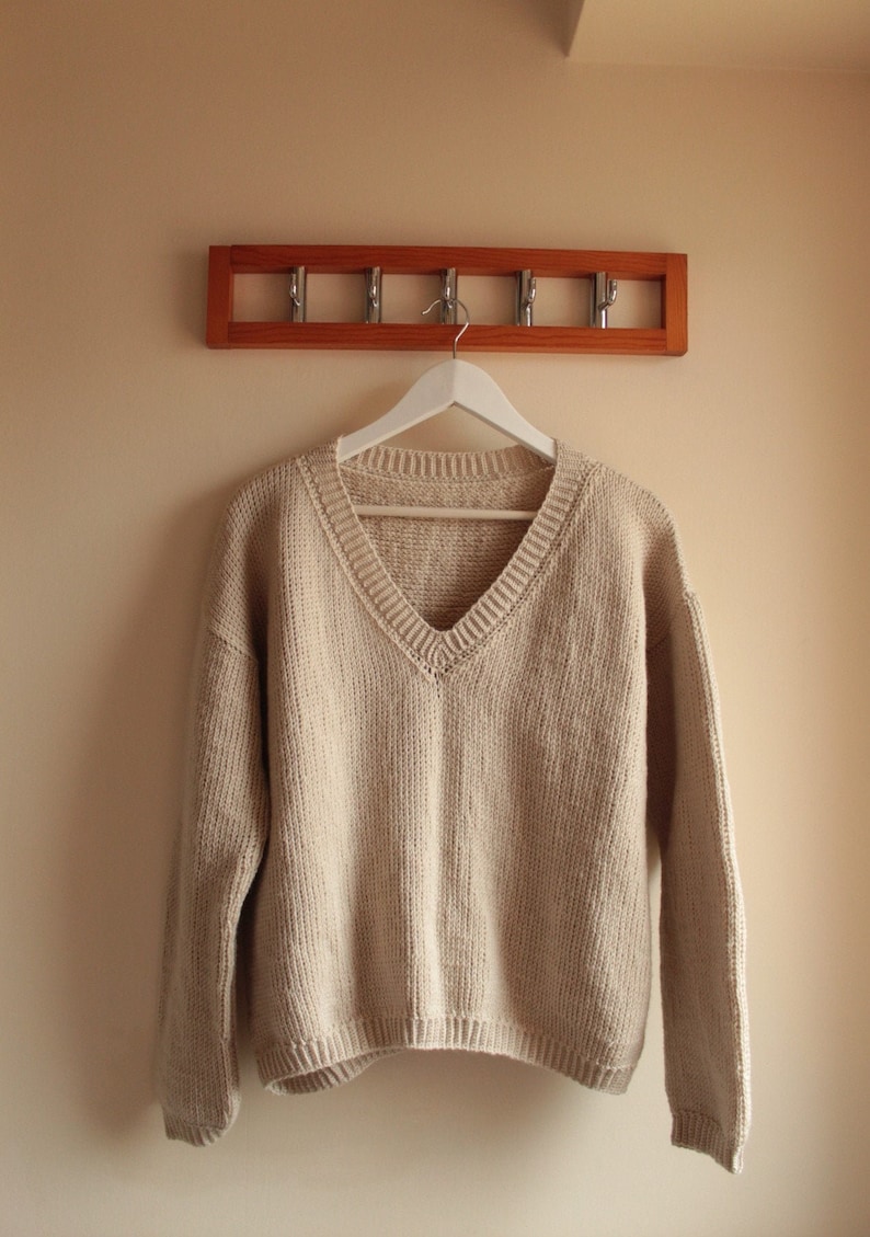 A beige, hand-crocheted V-neck sweater hanging on a white hanger, which is hooked onto a wooden wall-mounted coat rack with five metal hooks. The background wall is painted in a light cream color.
