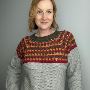 May include: A grey crochet sweater with a colour block design. The upper section features a pattern of red, orange, and green, with a dark green neckline. The sleeves and lower body are a solid grey colour.